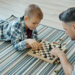 Father and son playing chess on a striped blanket.
